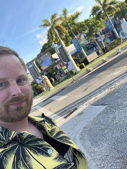 The image features a man wearing a yellow shirt with palm trees printed on it, standing on a sidewalk next to a street. The photo has been edited to include text that reads "GM Have a great start of your week And don' t forget to use @solflare". This suggests that the photo was taken as part of a social media post or promotional material for a business or event related to palm trees, given the man's shirt and the presence of palm trees in the image.