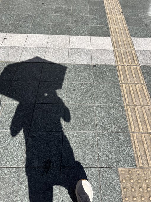 The shadow of a person holding a square umbrella, with another shadow and a white sneaker visible on a tiled sidewalk with tactile paving.