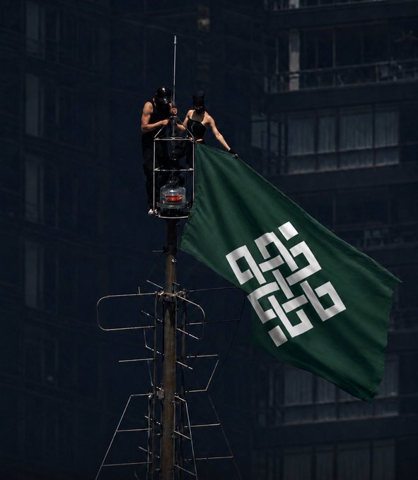 Two people in black masks stand on a rooftop antenna structure hoisting a large green flag bearing a white geometric logo.