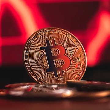 A physical Bitcoin (BTC) coin stands upright against a red-lit background, with additional coins scattered in the foreground. The image relates to the post by visually representing the cryptocurrency at the center of the bearish price forecast predicting BTC could drop to $48,000.