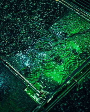 An extreme close-up of water droplets on a green-tinted glass surface, creating a textured, abstract pattern of circular ripples and light reflections.