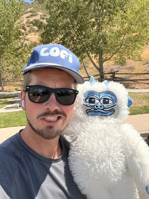 A man wearing a blue 'COFi' cap and sunglasses takes a selfie outdoors, posing next to a white fluffy Yeti plush toy.