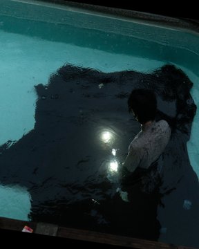 A dark, moody photograph of a person with long dark hair in a white shirt standing in a swimming pool at night, illuminated by a single light reflecting on the water's surface.