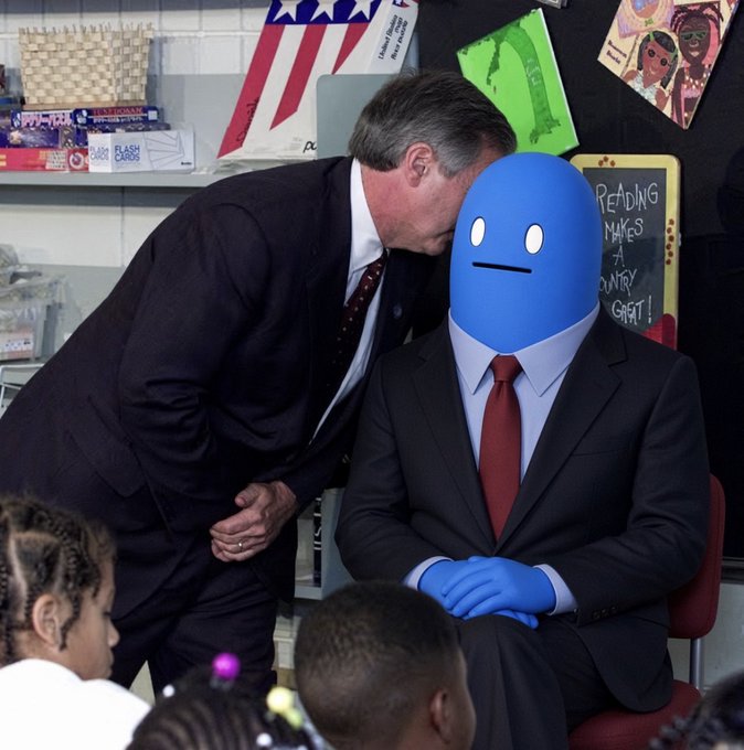 A man in a suit leans in to whisper to another seated man in a suit, whose face and hands are obscured by blue cartoon-style graphics, in what appears to be a classroom setting.
