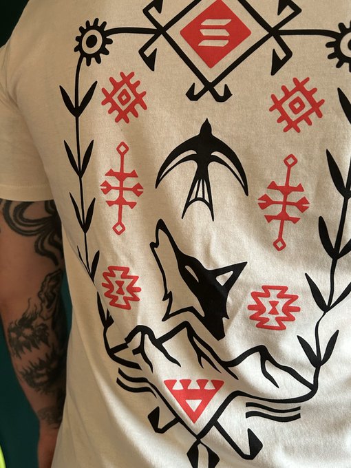 A person wearing a cream-colored t-shirt with a graphic print featuring a black wolf's head, a fox-like creature, red geometric symbols, and stylized plants; the wearer's tattooed arm is visible on the left.