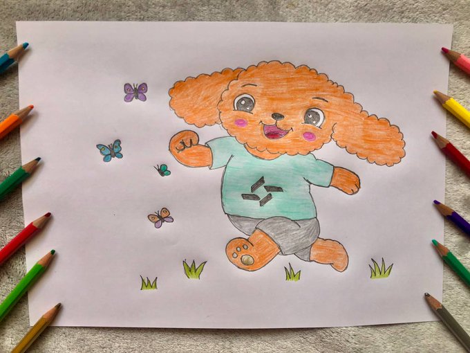 A child's hand-drawn illustration of an orange puppy wearing a teal shirt with a logo, surrounded by butterflies and grass, sitting on paper surrounded by colored pencils.