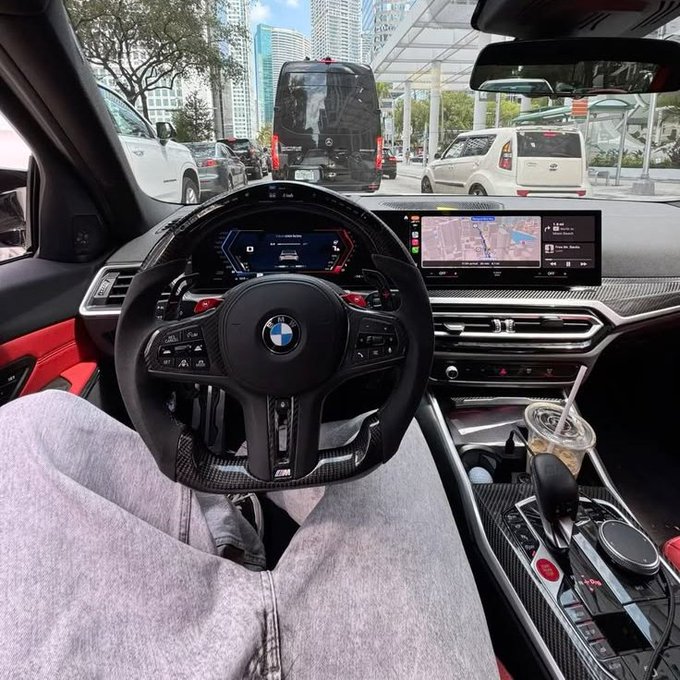 View from the driver's seat of a BMW (likely an M3) with a digital dashboard and navigation screen, facing a black Mercedes Sprinter van and city traffic ahead. Captioning "GM to whoever sees this" captures the casual, everyday greeting shared with strangers stuck in the same urban commute.