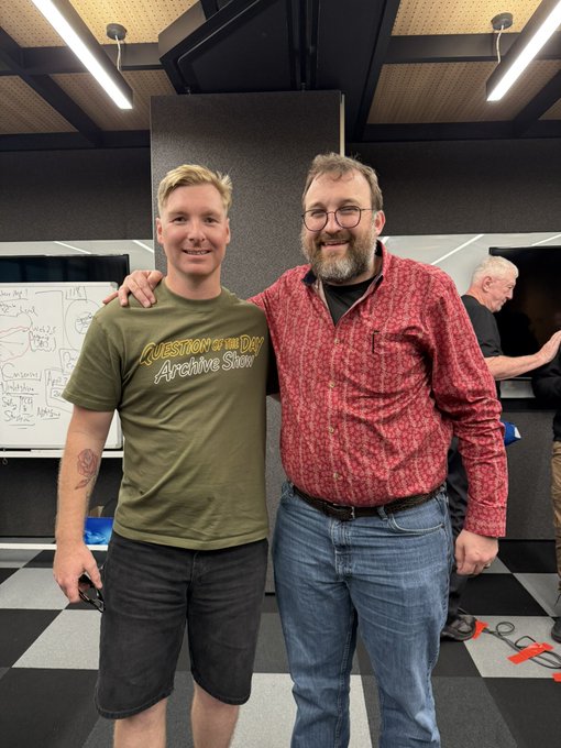 Two men pose together indoors; the younger man on the left wears an olive green 'Question of the Day Archive Show' t-shirt and dark shorts, while the older bearded man on the right wears a red patterned button-down shirt and jeans, with his arm around the younger man's shoulder.