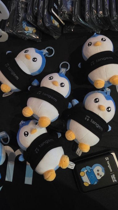 Several plush penguin keychains wearing black "tangem" hoodies are displayed on a dark surface, accompanied by a matching card case and packaged items in the background.