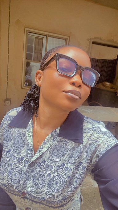 Selfie of a woman wearing large dark sunglasses and a blue patterned blouse with a dark collar, standing outdoors near a building.