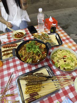 The image shows a dining table with various plates of food, including a large bowl filled with noodles, several bowls containing different dishes, and an assortment of skewers. A person can be seen sitting at the table, likely enjoying their meal or preparing to eat. The table is covered in a checkered tablecloth, adding a touch of elegance to the scene. 

In addition to the food items on the plates, there are also several bottles and cups scattered around the table, suggesting that beverages have been served alongside the dishes. A cell phone can be spotted near one of the bowls, possibly used for capturing memories or communicating with others during this mealtime experience.