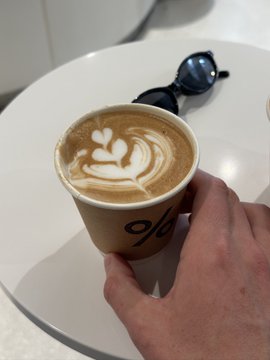 A hand holds a latte with heart-shaped latte art in a paper cup marked "%", with sunglasses resting on a white table in the background—pairing a coffee break with a casual ask about favorite coins to stake on Tangem.