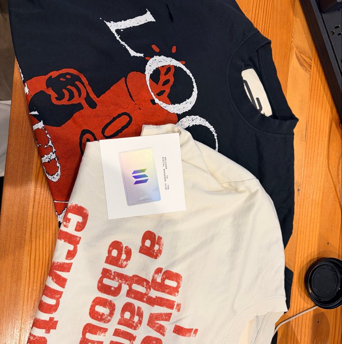 Two folded shirts—one black and one white—rest on a wooden surface, both featuring red and white printed graphics including a stylized figure, circular motifs, and the repeated text "MORE MORE" and "THE MORE." A holographic Solana ("SOL") card sits atop the white shirt, tying the apparel display to the "solana summer kit" merchandise bundle.