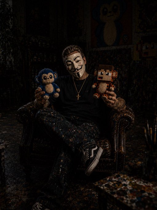 A person wearing a V for Vendetta Guy Fawkes mask and black clothing sits in a dark leather armchair holding a small blue stuffed monkey and a pixelated Minecraft-style plush toy, with paint-splattered pants and a cross necklace.