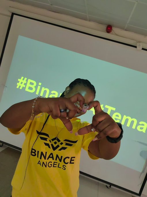 Person wearing a yellow 'BINANCE ANGELS' t-shirt and headphones, posing with hands forming a heart/frame shape, with a projection screen showing '#Binance...' in the background.
