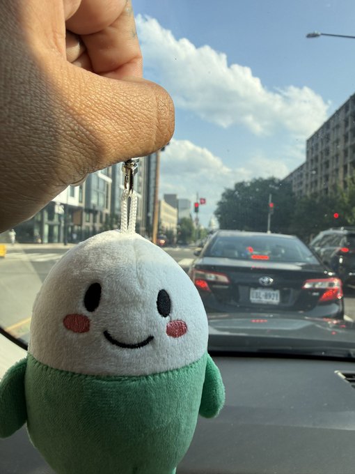 A hand holding a green and white plush keychain character dangling in front of a car windshield showing a street scene.