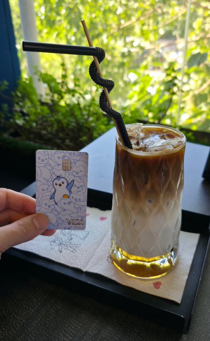 A hand holds a blue Pokemon-themed Visa card next to a layered iced coffee with a uniquely knotted straw.