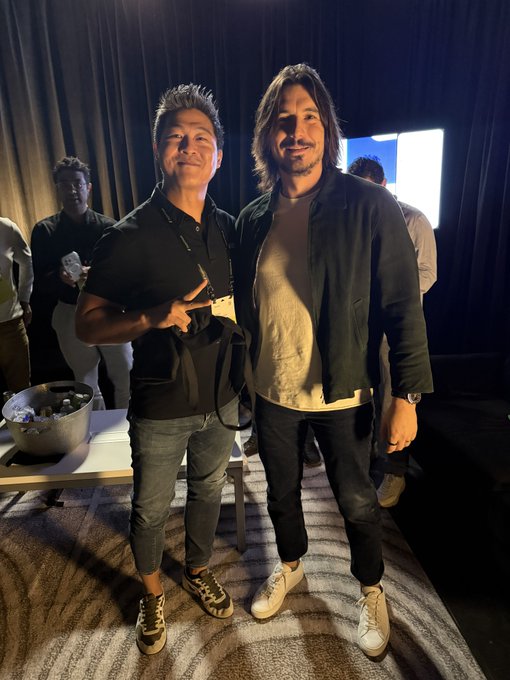 Two men pose together at what appears to be a social event; one wears a black polo shirt with a lanyard and the other wears a black jacket over a white shirt.
