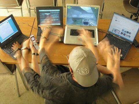 A man wearing a baseball cap sits at a wooden desk juggling four open laptops simultaneously, with multiple hands visible as he types, surrounded by cables and a cell phone.