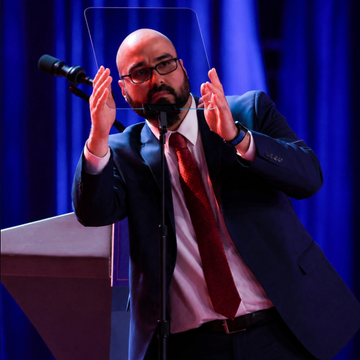 A bald man with glasses and a beard, wearing a dark suit and red tie, stands at a podium gesturing with both hands raised while speaking into a microphone, behind a clear teleprompter. The image illustrates the news that Gabriel Perez, Trump's teleprompter operator, was fined $172,000 by the CFTC for betting on the president's speeches through Kalshi trades.