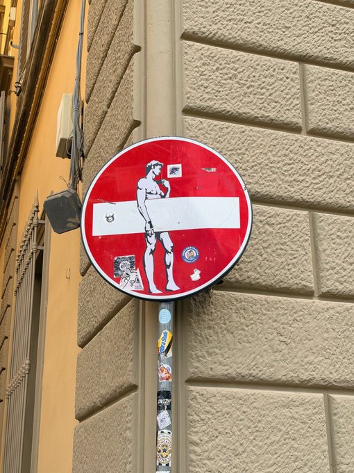 A red and white 'Do Not Enter' street sign in Italy has been modified with black-and-white illustrations of a naked statue of David on the man, with stickers plastered on the pole and sign below.