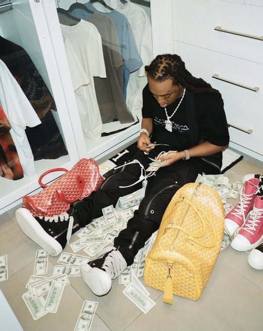 A person with braided hair wearing a black t-shirt and chain, sitting on a floor covered with stacks of cash, surrounded by designer clothing, a pink crocodile-patterned bag, and sneakers.