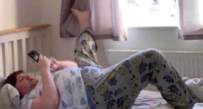 A man in pajamas lies on a bed scrolling on his phone, with one foot propped up against the curtains.