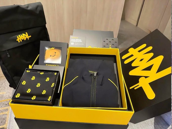 A "Binance TH" branded yellow and black swag box containing a folded hoodie, a gift-wrapped item, and a card, accompanied by a black backpack featuring the same logo. The branded merchandise directly reflects the post's gratitude toward the Binance TH team for the thoughtful gift and community support.