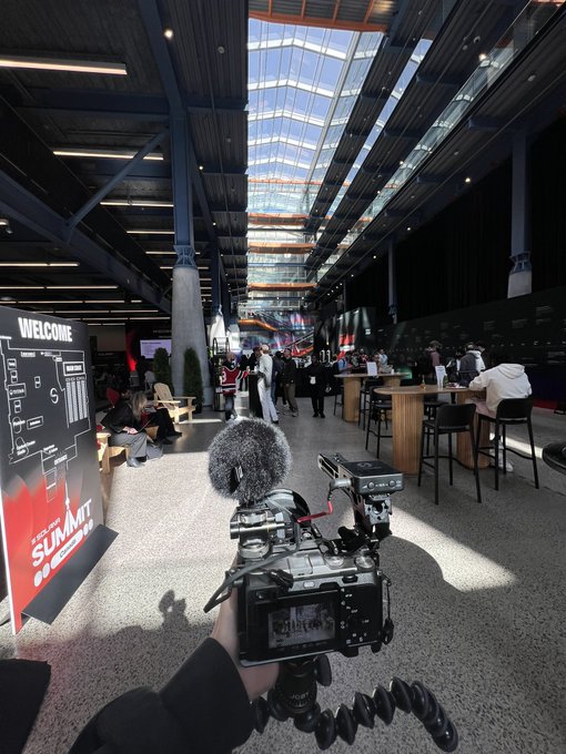 A first-person view shows a hand holding a camera with a fluffy microphone on a flexible tripod, pointing toward a large, modern indoor venue with a glass ceiling and a 'WELCOME' sign reading 'YIP SUMMIT'.