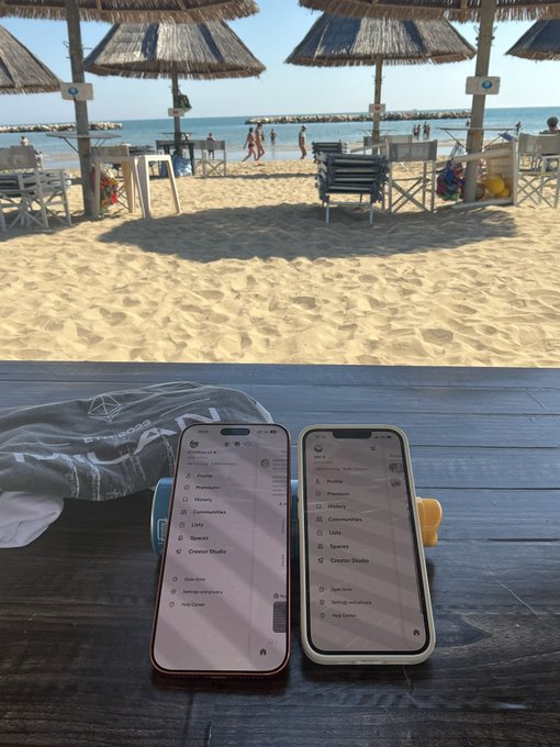 Two smartphones displaying social media menu screens sit on a wooden table at a beach, with thatched umbrellas, sand, and the sea visible in the background.