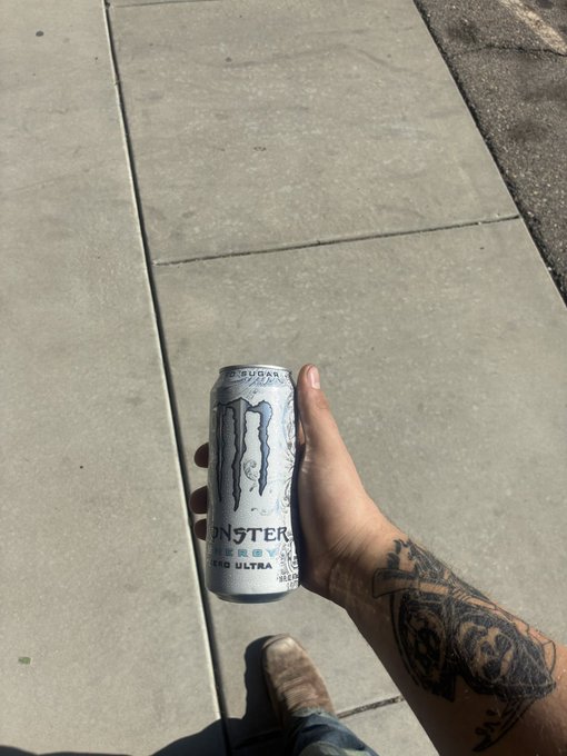 A tattooed hand holding a Monster Energy Zero Ultra sugar-free can on a sunlit sidewalk.