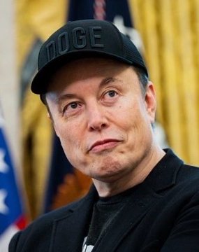 Photo of Elon Musk wearing a black cap with 'DOGE' printed on it, standing in front of American flags.