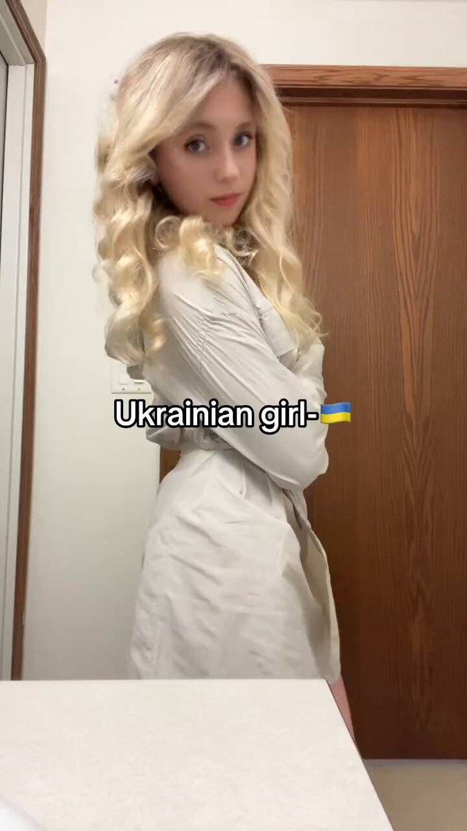 The image features a young woman with blonde hair, dressed in a white dress, standing against a wooden wall. She appears to be posing for a photo or video, possibly sharing her outfit choice with others. The background of the image includes text that reads " Ukrainian girl - $BTC", which might indicate an online store selling clothing items from Ukraine.