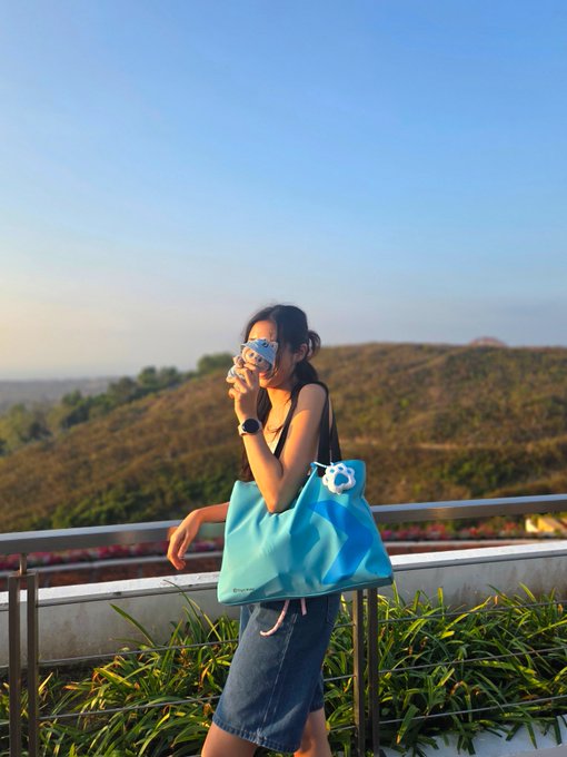 A young woman poses on a balcony during golden hour, sipping from a blue-patterned cup while carrying a bright blue tote bag, with rolling hills bathed in warm sunset light behind her—visually echoing the post's "golden hour" optimism about crypto surpassing $3T. The sun-kissed, upward-looking scene mirrors the bullish, triumphant mood of the milestone announcement.