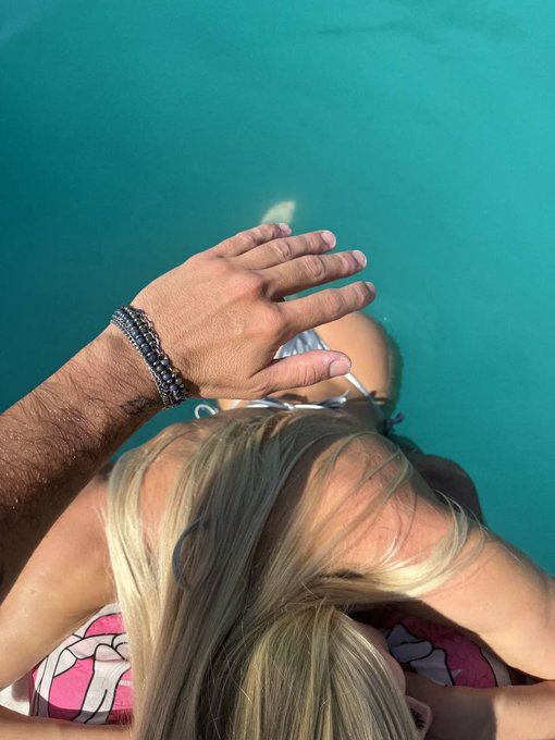 A hand with a beaded bracelet rests on the shoulder of a person in a pool, with their blonde hair floating in the water, creating a relaxed "gm" vibe.