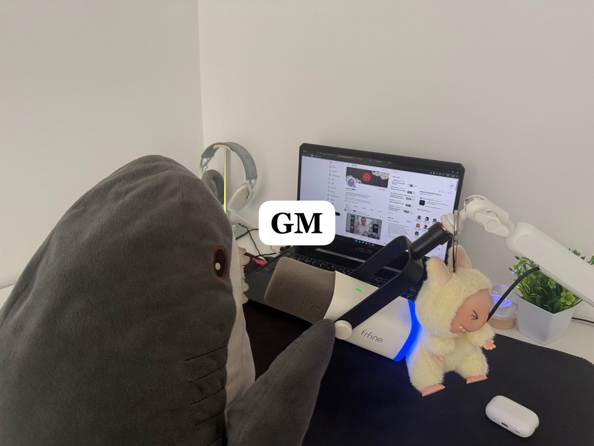 An over-the-shoulder view of a person in a gray shark onesie sitting at a desk with a laptop, an Fifine microphone, headphones, and a Labubu keychain; a 'GM' text sticker is overlaid in the center.