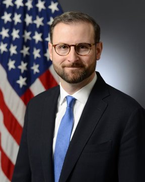 Official portrait of a man with glasses and a beard wearing a dark suit and blue tie, standing in front of an American flag.