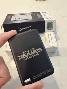 A hand holds a black Ledger device with 'MoonPay xGAMES LEAGUE' printed on it, with a Ledger product box visible in the background.