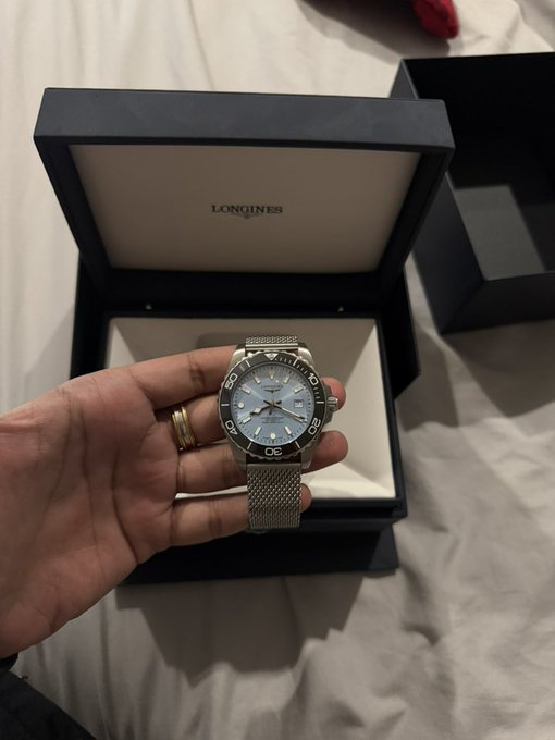 A hand wearing a gold ring holds a Longines watch with a light blue dial and steel mesh band in front of its open Longines presentation box on a white bed.