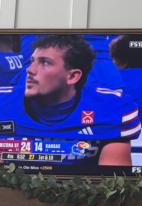 A photo of a TV screen showing a Florida Gators football game on FS1, with Arizona State trailing Kansas 24-14 in the 4th quarter (6:52 remaining); a young man in a blue Florida jersey sits on the bench looking up.