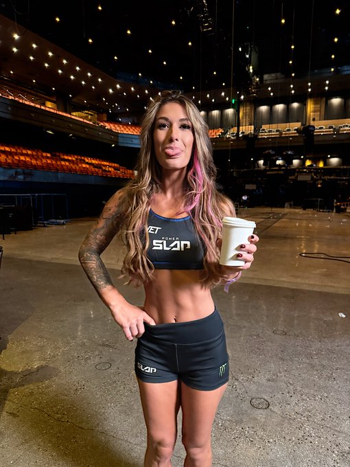 A woman with long brown hair featuring pink highlights poses in an empty arena wearing a 'SLAP' branded sports bra and shorts, holding a white coffee cup, with visible tattoos on her arm.