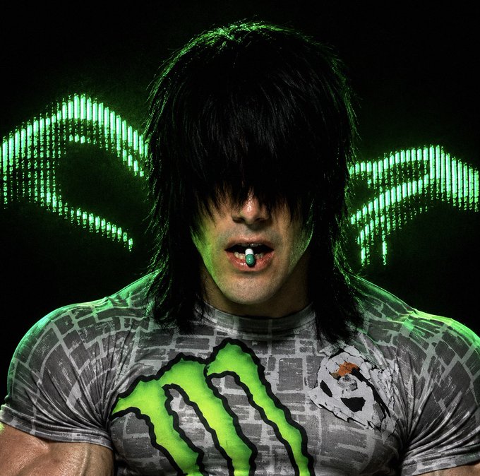 A muscular man with long dark hair wearing a green Monster Energy shirt, photographed against a background with green lit wings and a full moon.