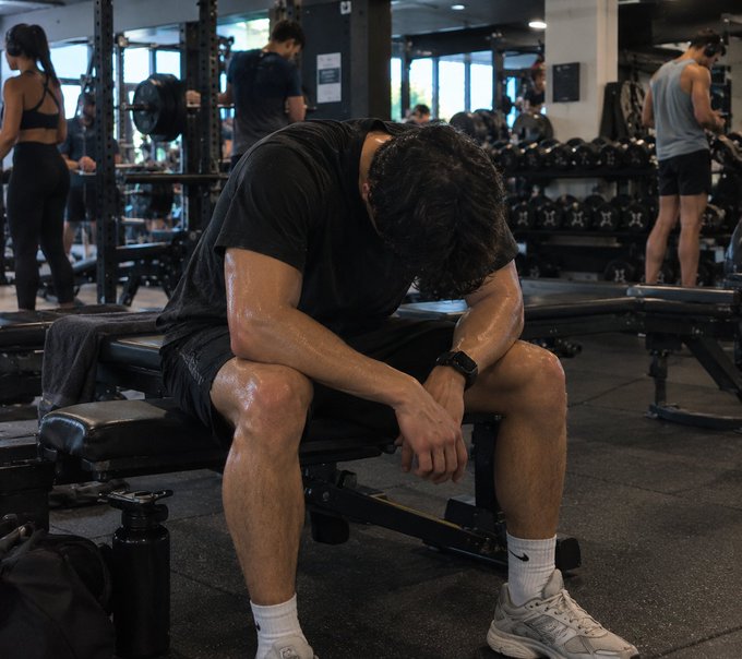 A gym-goer sits hunched over on a weight bench, head down and visibly exhausted—mirroring the "wiped out" sentiment of crypto traders after nearly $1 billion in liquidations on Oct 10, 2026.
