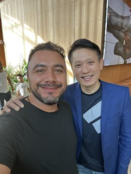 Two men smiling in a selfie, one in a black shirt with a beard and the other in a blue blazer over a gray t-shirt, appearing to pose together in a brightly lit room.