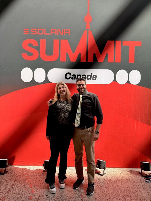 Two people pose together in front of a red "SOLANA SUMMIT Canada" backdrop, smiling for the camera. The setup appears to be a stage area, fitting the post celebrating their co-MCing duties on Day 2 of the event.