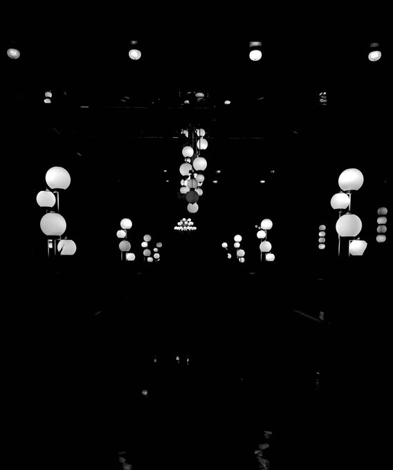 A high-contrast, black-and-white photograph of an interior space illuminated by numerous spherical white lights hanging at various heights against a dark background.