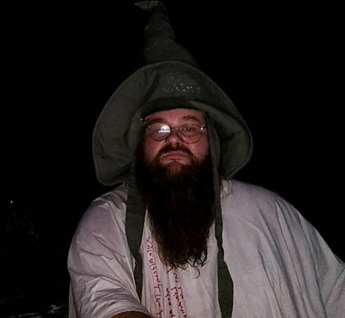A man with a long dark beard, glasses, and a tall pointed grey wizard hat, wearing a light-colored robe, photographed at night.