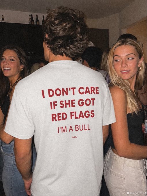 A man at a party wears a white t-shirt reading "I DON'T CARE IF SHE GOT RED FLAGS I'M A BULL" in red text, standing between two women — the shirt's defiant bullish attitude perfectly mirrors the post's caption about ignoring warnings and staying bullish after a losing $BTC trade.