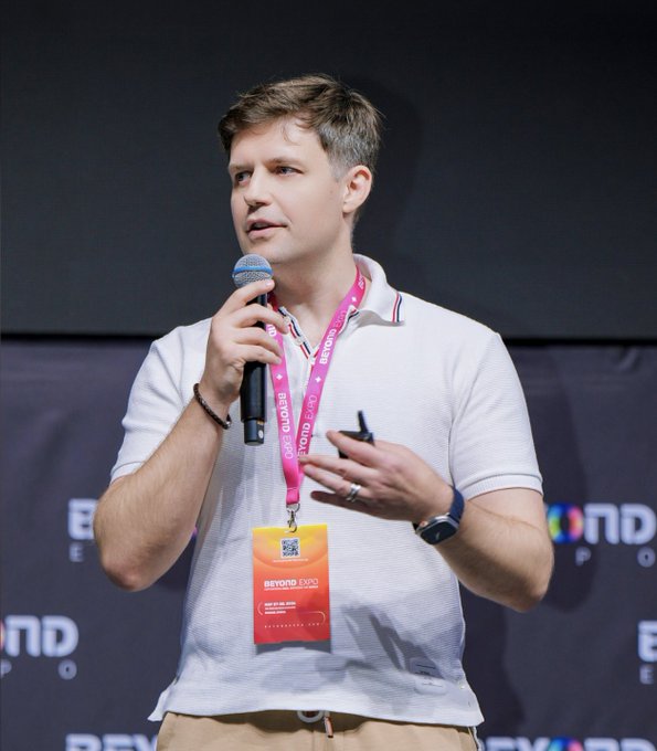 The image features a man standing on a stage at an event, holding a microphone and wearing a white shirt with a pink lanyard around his neck. The background of the image shows a black screen displaying text related to AI research and development. The caption for the image reads "At @beyondtechexpo in Macao, @michaelh_0g put the 0G thesis on the main stage: AI needs a trust layer. Private inference, verifiable compute, storage and coordination have to sit inside the infrastructure layer."