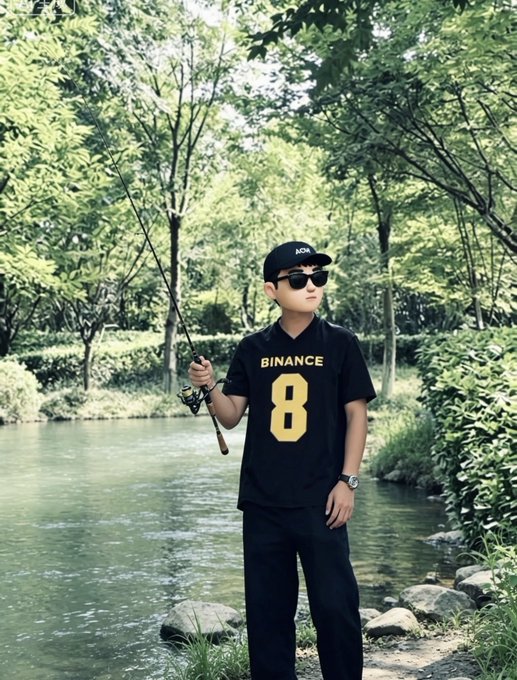 Young boy wearing a black 'BINANCE' jersey with the number 8, sunglasses, and a black cap, standing by a river in a wooded area while holding a fishing rod.